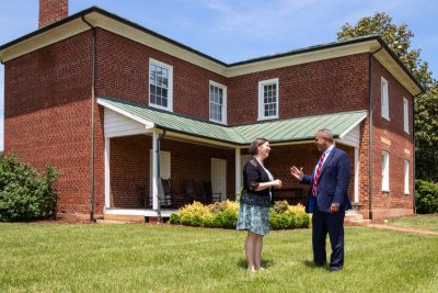 A man and a woman walk in the green lawn outside a large brick home.