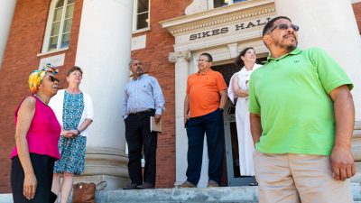 A group of people stand on the steps of a big brick building under a sign that reads "Sikes Hall."