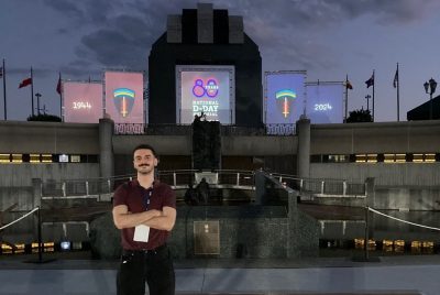 A man stands with crossed arms in front of tiers of concrete with steps, leading towards three movie screens in the distance with the words “80 YEARS NATIONAL D-DAY MEMORIAL.”