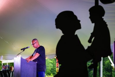 A black silhouette cutout in the foreground with a man standing at a podium, head lowered in thought, in the background.