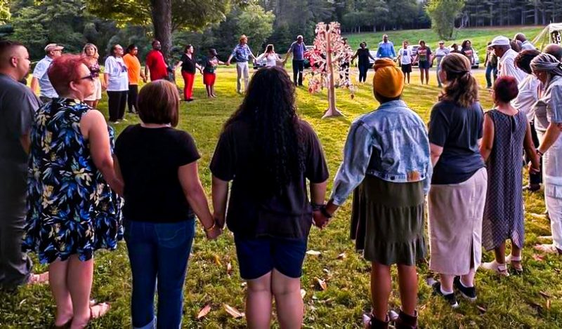 About 40 people stand in a circle holding hands with a fake tree in the center.