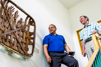 Two men stand at the top of a stairway looking at an art piece made from an antique basket and sculptured clay hands.